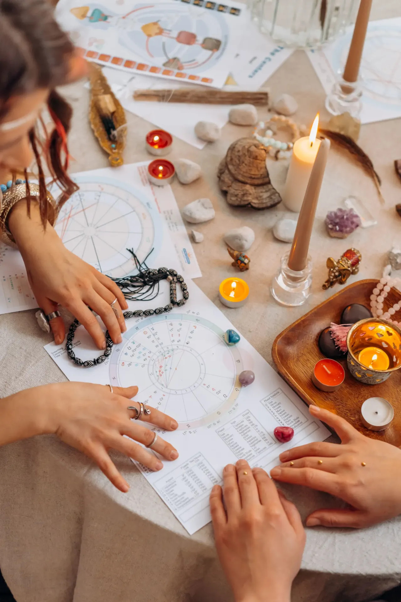 Mâini așezate pe masă cu lumânări aprinse, cristale și hărți astrologice, în timpul unei ședințe.