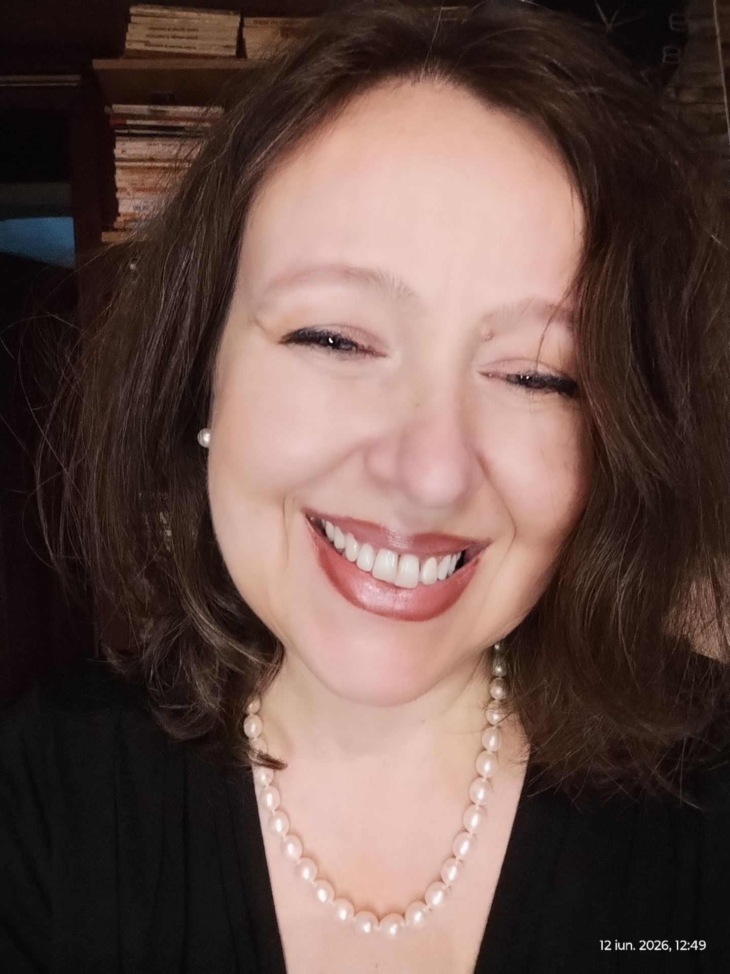 Merie-Luisa Beldiman zâmbitoare, cu păr brun si ochi albastri, colier de perle și fundal cu rafturi.de carti